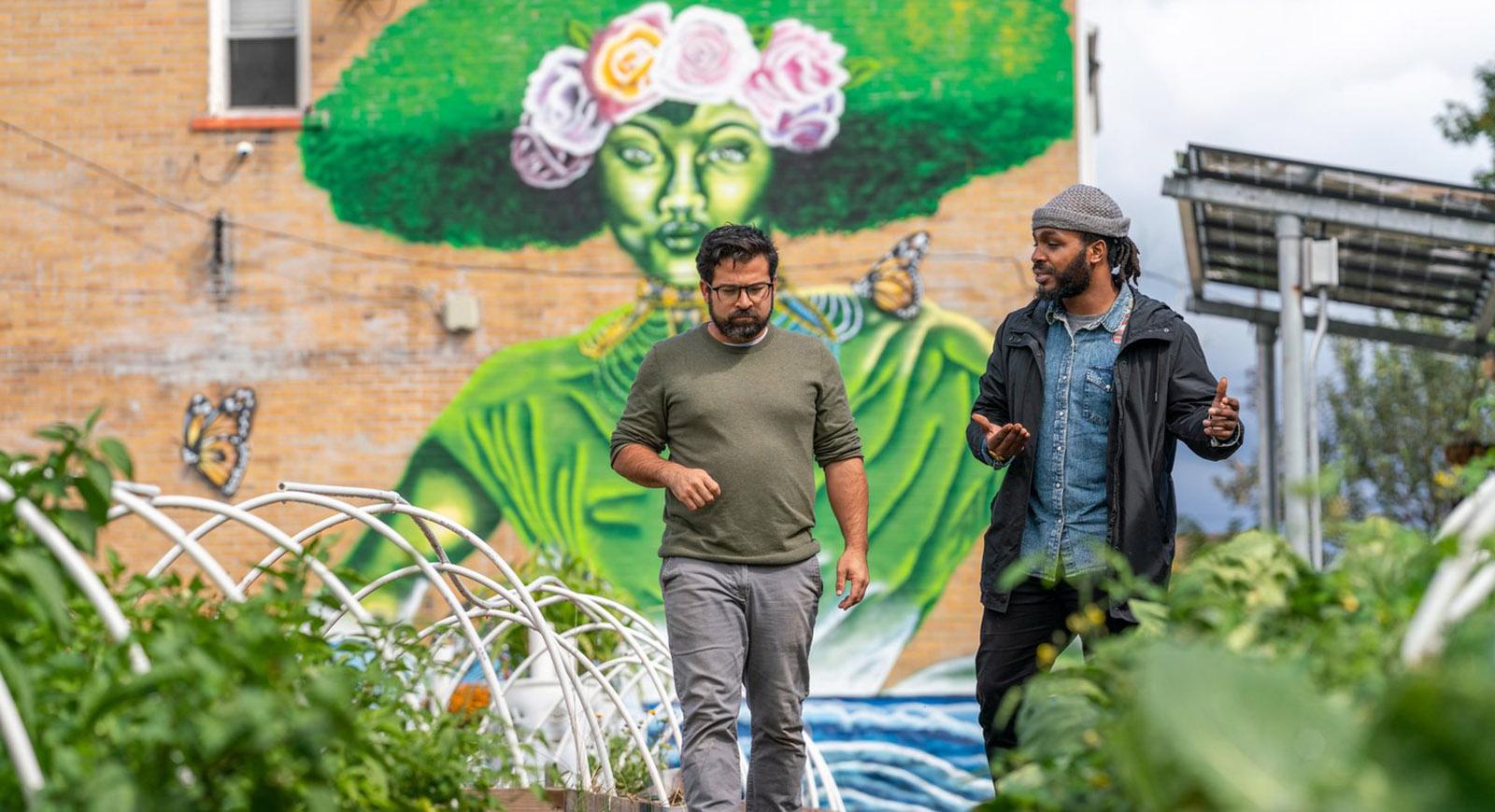 Photo of two men walking and talking in a community garden, with a brick wall with a large and colorful mural in the background